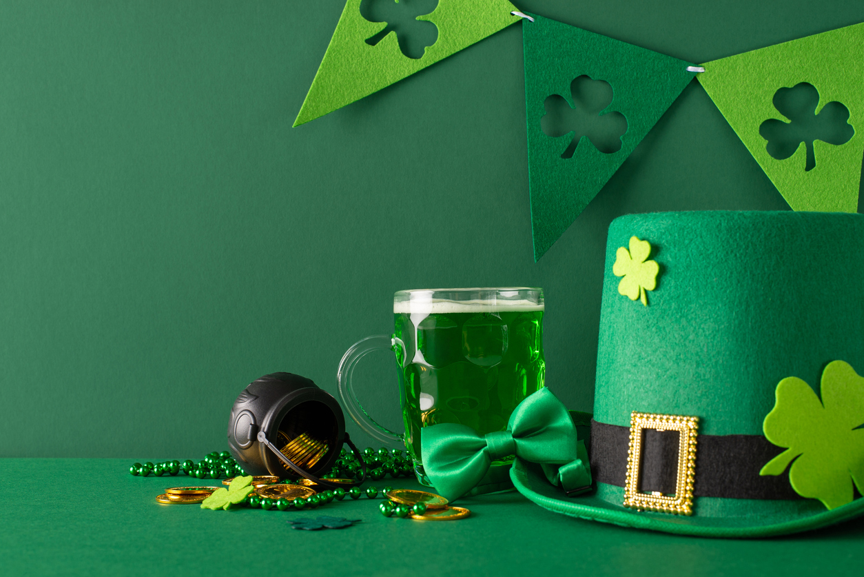 St. Patrick's Day theme: side view shot featuring pint of beer, green shamrocks, glittering gold coins, mystical pot, leprechaun hat, bow tie, beads, flag garland, all set on vibrant green backdrop