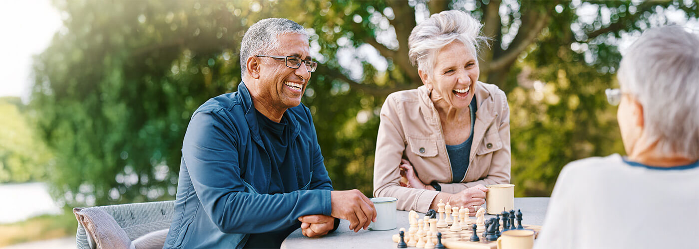 Senior man and woman sit at an outdoor table playing chess while smiling