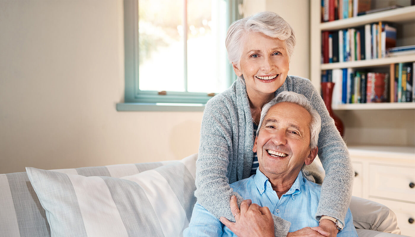 happy-senior-couple-embracing-couch Smiling senior man sits on couch while smiling senior woman stands behind and embraces him